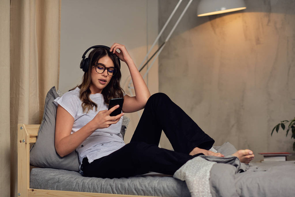 Pretty Caucasian brunette in pajamas and with headphones on head using smart phone late at night while sitting on the bed in bedroom. - Фото, зображення