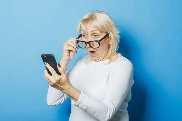 Gözlüklü şaşırmış bir yüzü olan yaşlı kadın mavi bir arka plan üzerinde telefona bakar. Konsept Sms bildirimleri, yeni teknolojiler, promosyon ve satış, bildirim - Fotoğraf, Görsel