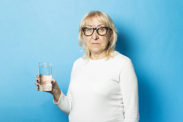 Velha senhora com óculos segurando um copo de água contra um fundo azul. Conceito de sede, desidratação
 - Foto, Imagem