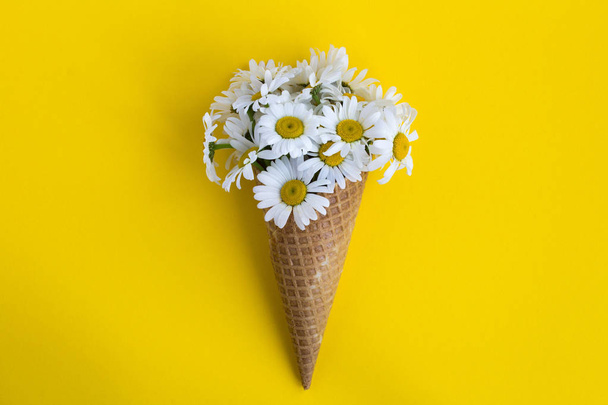 Ice cream cone with chamomiles  in the center of the  yellow background.Top view.Copy space.Spring or summer flowers concept. - Фото, изображение