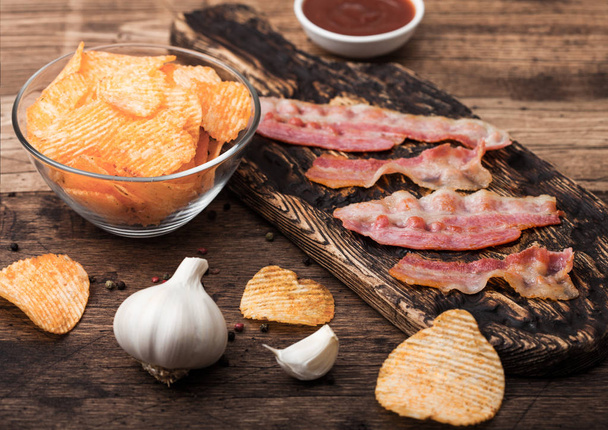 Potato crisps with backon flavour with grilled bacon rashers on vintage chopping board with garlic and sauce on wooden background. - Φωτογραφία, εικόνα