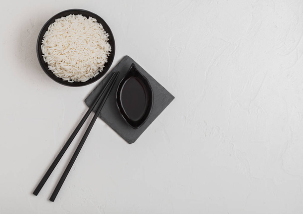 Black bowl with boiled organic basmati jasmine rice with black chopsticks and sweet soy sauce on stone mat on white background. Space for text - Photo, image