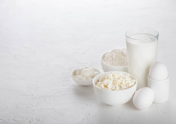 Fresh dairy products on white table background. Glass of milk, bowl of baking flour, cottage cheese and mozzarella. Eggs and cheese. Space for text - Photo, Image