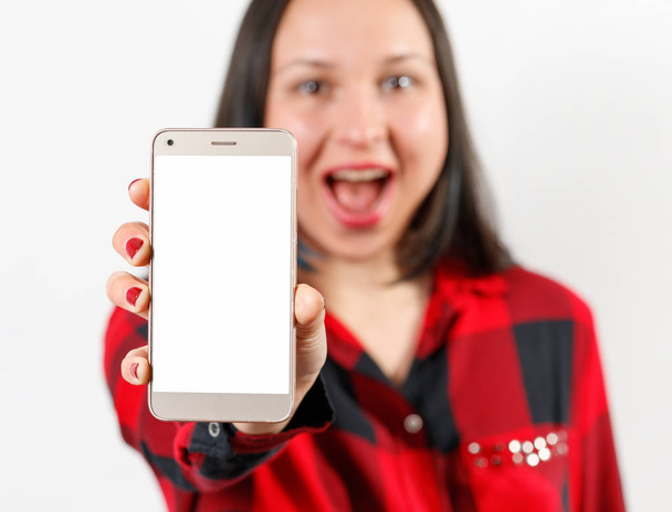 a young girl woman in a red and black shirt is holding a smartphone with a blank white screen vertically in front of her - Foto, Imagen