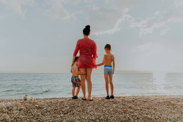 Τα παιδιά στέκονται στην παραλία μαζί με τη μητέρα τους κοιτάζοντας το ηλιοβασίλεμα. - Φωτογραφία, εικόνα