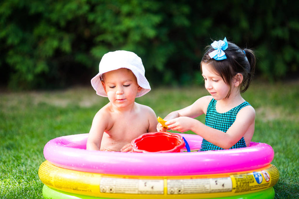 Thema Kindersommerspiele im Hof. Kaukasisches Geschwisterpaar spielt Plastik-Spielzeug-Eimer im Wasser, aufblasbares rundes Kinderbecken. Sommer ist heiß, Ruhe in Badeanzügen - Foto, Bild