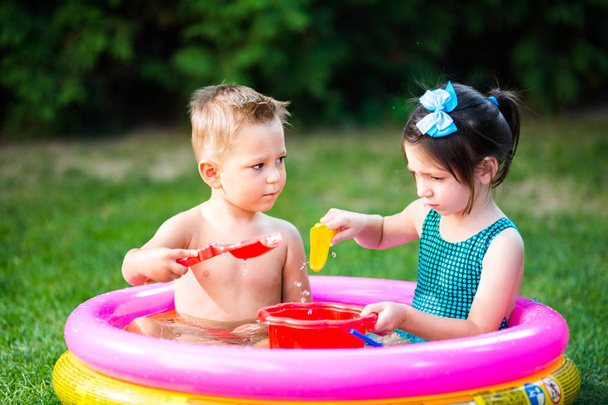Subject childhood summer games in the yard. Caucasian brother and sister playing plastic toys bucket sitting in the water, inflatable round children's pool. Summer is hot, rest in swimsuits - Photo, Image