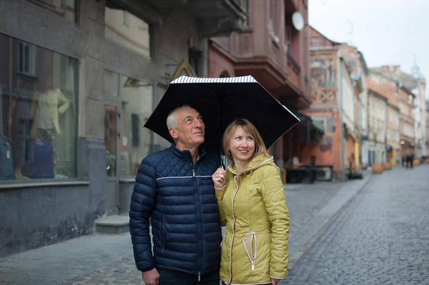 Outdoors portrait of elderly man and his young blonde-haired wife embracing each other standing under their umbrella on paved street. Couple with age difference. - Photo, Image