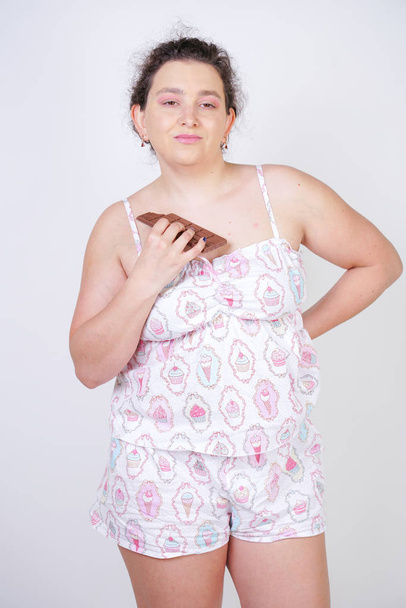 bochtige vrouw in grappige pyjama met een chocoladereep staat op een witte achtergrond in de Studio - Foto, afbeelding