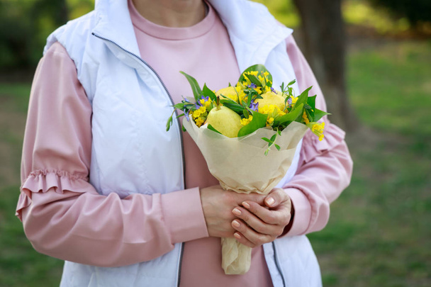Cute bouquet of flowers and fruits in the hands of a young woman on a background of green grass - Foto, Bild