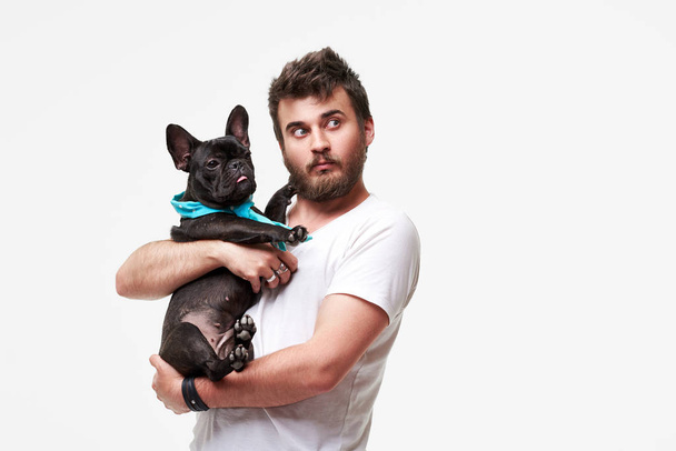 Hipster vousatý chlapík, který držel v náručí hezkého francouzského Bulldoga s láskou a pohrává si s ním, na šedém pozadí - Fotografie, Obrázek