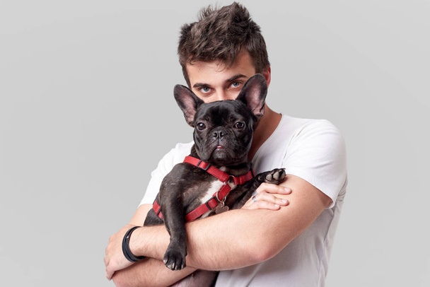 Hipster Bearded guy holding and hugging a nice French Bulldog dog in his arms with love and playing with him, against a grey background - Photo, Image