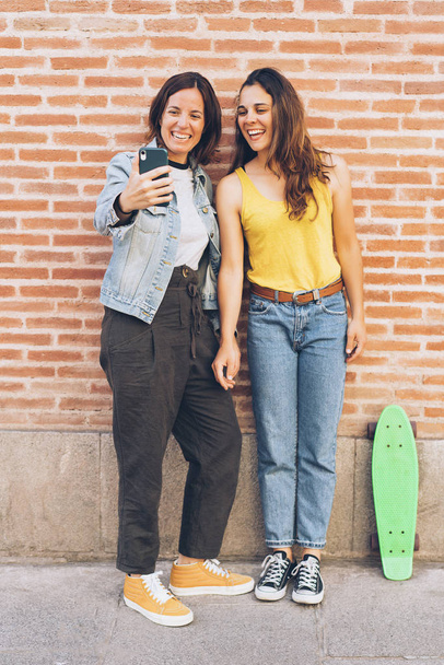 Young women couple making a selfie. Behind brick wall. Positive emotion and tolerance concept. - Photo, image