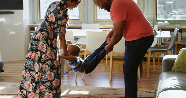 Vista laterale dei genitori afroamericani che dondolano il figlio in una casa confortevole. Sorridono e si divertono. 4k
 - Filmati, video