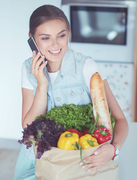 Smiling woman with mobile phone holding shopping bag in kitchen - Foto, immagini