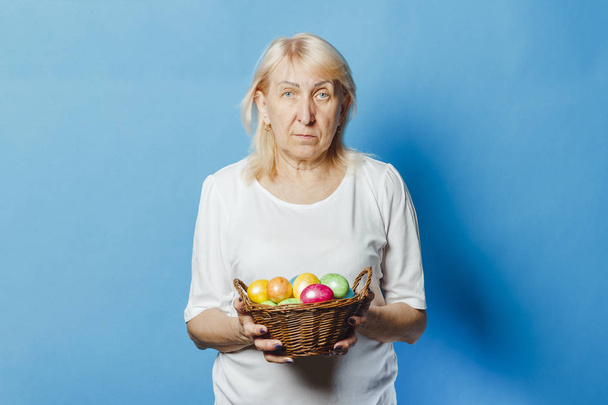 La anciana sostiene en sus manos una canasta de huevos de Pascua conmigo sobre un fondo azul. Concepto de celebración Pascua
 - Foto, Imagen