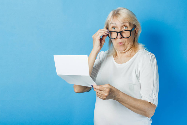 La anciana sostiene en sus manos una hoja blanca y limpia de papel sobre un fondo azul. El concepto de recibir cartas, notificaciones, facturas de pago, impuestos
 - Foto, imagen