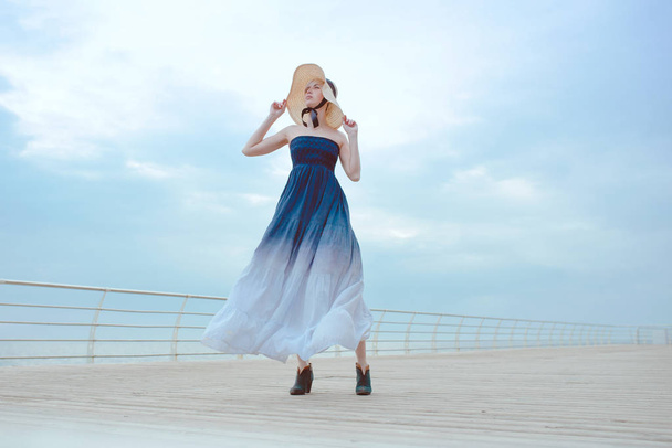 Caucasian beautiful stylish blonde sad woman in blue dress and in straw hat standing by the sea  - Zdjęcie, obraz