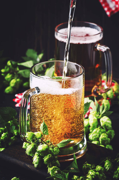 Large mugs with german light beer and fresh hops, pours into glass, dark table, selective focus - Foto, Imagem