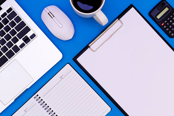 Plochá tabulka kancelářského stolu moderního pracoviště s notebookem na modrém stole, špičatý pohled na notebook a prostor pro kopírování na černém pozadí, modrá kancelářská kancelář s notebookem, smartphone a další pracovní potřeby s šálkem kávy. - Fotografie, Obrázek