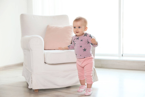 Cute baby girl standing near armchair in room - Foto, immagini