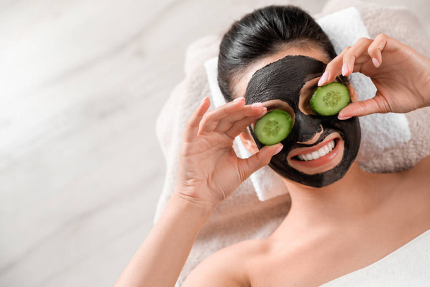 Beautiful woman with black mask on face and cucumber slices relaxing in spa salon, top view. Space for text - Photo, image