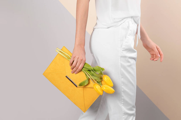 Stylish woman with clutch and spring flowers against color background, closeup - Фото, изображение