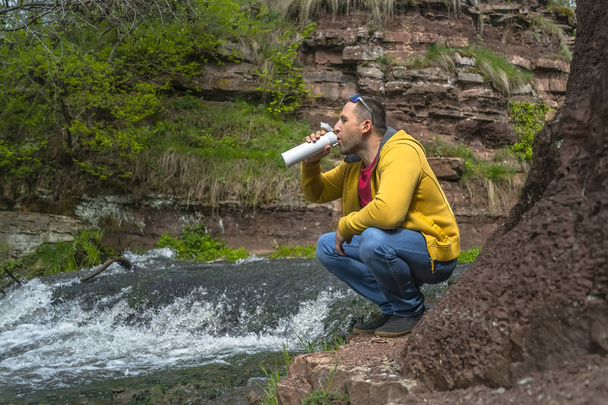 Man drinkt koffie van witte Thermos in de buurt van Mountain River - Foto, afbeelding