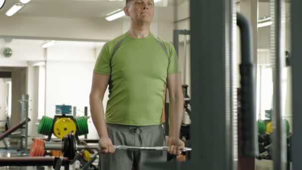 A overweight man lifts an ez barbell while standing at the gym. Exercise for biceps. Fitness. Healthy lifestyle. - Záběry, video