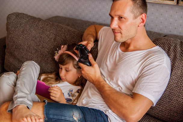 Little cute girl is using tablet, her father is playing console .   - Photo, Image