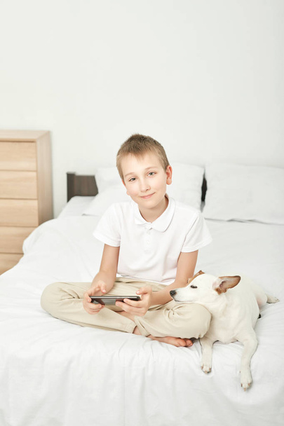 chłopiec z psem Jack Russell grając na smartfonie na białym łóżku w domu - Zdjęcie, obraz