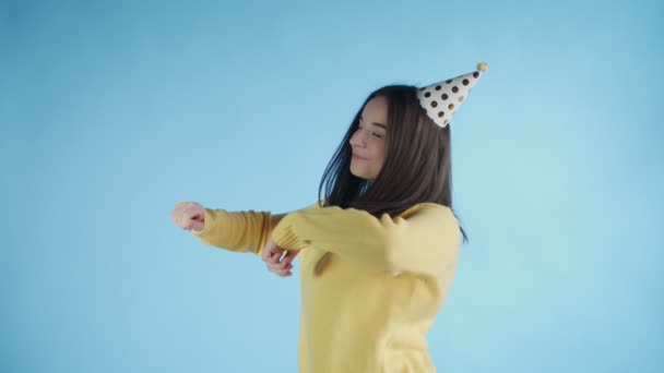 Young woman in yellow sweater with party hat is dancing on a blue background - Imágenes, Vídeo