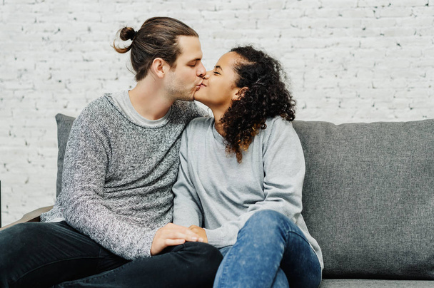 Porträt eines jungen multiethnischen Paares, das sich zu Hause auf der Couch küsst - Foto, Bild