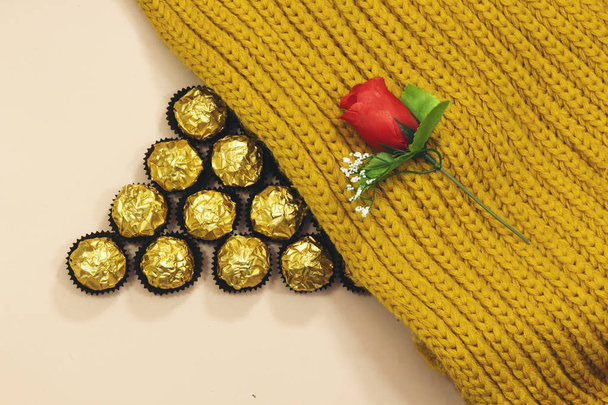vytvoření trojúhelníku od zlatých prémiových čokoládových sladkostí pod vlněné zimní šátkem a červenou květinou - Fotografie, Obrázek