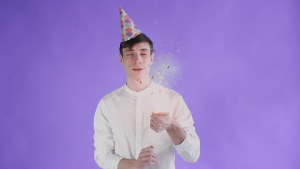 Young man exploding confetti cracker on a purple background. - Filmati, video