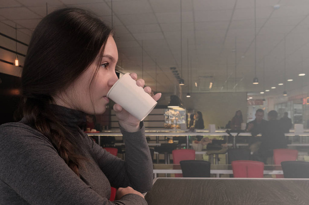 Νεαρή γυναίκα πίνει καφέ σε ένα καφέ. Το κορίτσι ζεσταίνεται μια φθινοπωρινή μέρα με ένα ζεστό ποτό. Περπατήστε στην εταιρεία ή μόνοι. - Φωτογραφία, εικόνα