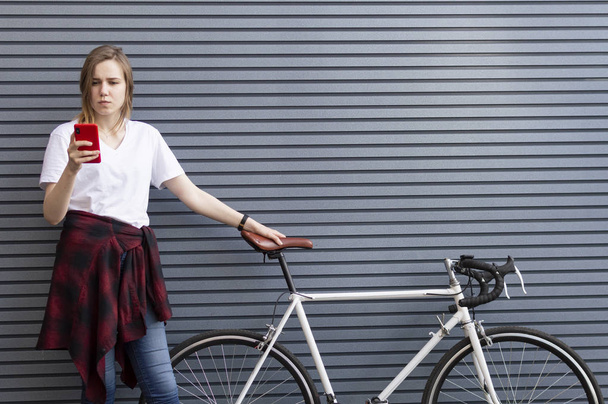 νεαρή όμορφη κοπέλα στέκεται με ένα ποδήλατο στο φόντο ενός τοίχου και κοιτάζει στο τηλέφωνο, μια γυναίκα ξεκουράζεται από ένα ταξίδι - Φωτογραφία, εικόνα