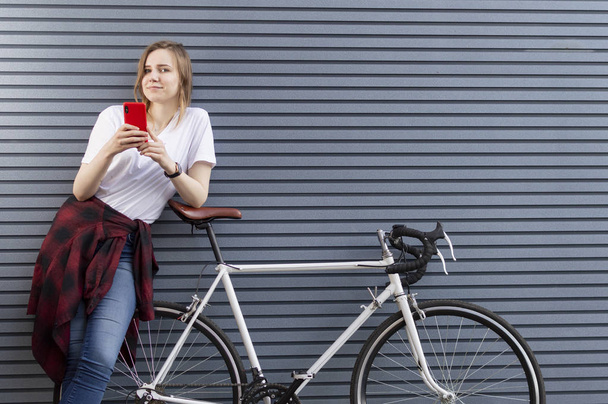 jeune belle fille se tient avec un vélo sur le fond d'un mur et regarde dans le téléphone, une femme utilise un smartphone et sourit
 - Photo, image