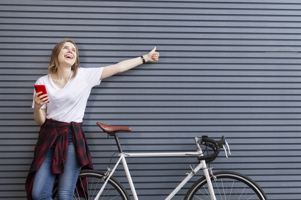 young beautiful girl stands with a bicycle on the background of a wall and looks into the phone, a woman shows a finger to a place for text - Φωτογραφία, εικόνα