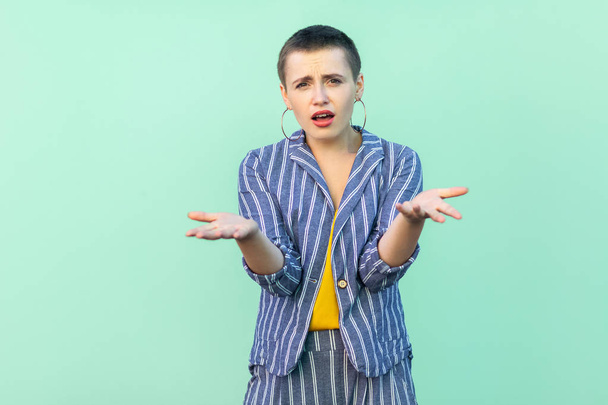 confused beautiful short hair stylish woman in casual striped suit standing with raised arms and looking at camera on light green background - 写真・画像