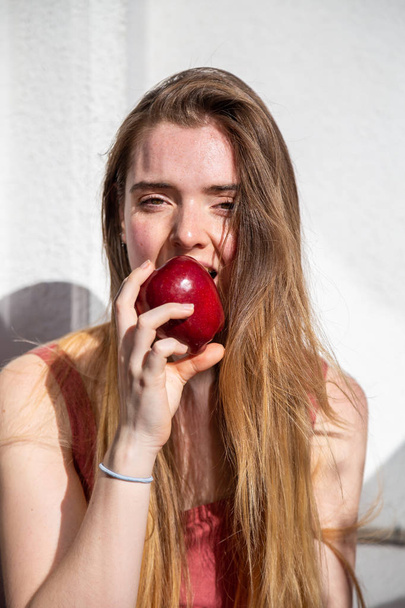 Молода радісна приваблива жінка в повсякденному літньому одязі сидить на балконі і їсть смачне соковите червоне яблуко
 - Фото, зображення