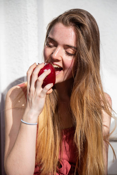 Young joyful attractive woman in casual summer dress sitting on balcony and eating tasty juicy red apple - Photo, image