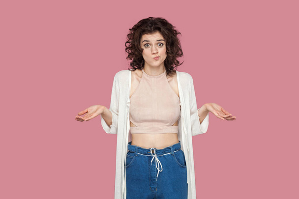 confused beautiful brunette young woman with curly hairstyle in casual style standing with raised arms and looking at camera on pink background - Fotó, kép
