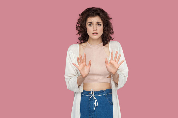 stressed scared beautiful brunette young woman with curly hairstyle in casual style looking at camera and showing stop sign on pink background - 写真・画像