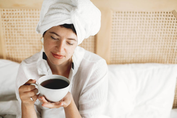 Όμορφη ευτυχισμένη νεαρή γυναίκα πίνοντας καφέ ή τσάι στο κρεβάτι σε ζεστό - Φωτογραφία, εικόνα