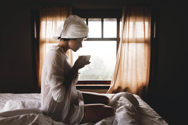 Gyönyörű boldog fiatal nő iszik kávét vagy teát az ágyban forró - Fotó, kép