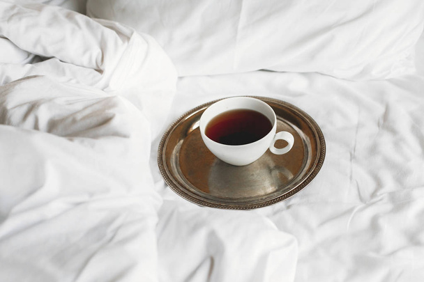 Tee in weißer Tasse auf metallischem Vintage-Tablett auf weißem Bett. Genießen - Foto, Bild