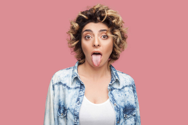 crazy funny young woman with curly hairstyle in casual blue shirt showing tongue out and looking at camera with big eyes on pink background - Photo, image