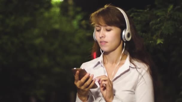 Veselá dívka cestuje v nočním městě. mladá dívka ve sluchátkách a smartphone poslouchá hudbu, tančí a usmívá se v noci v městském parku. Close-up - Záběry, video