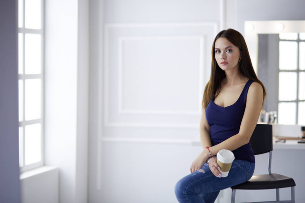Beautiful young woman holding coffee cup and looking away with smile while sitting on chair - Фото, зображення
